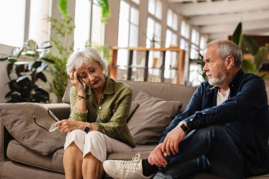 Oturma odasında hayal kırıklığına uğramış kocasının yanında otururken gözlük takan ve alnına dokunan yaşlı bir kadın ilişki sorunları ve iletişim sorunları yaşıyor.