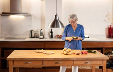Yaşlı kadın kabak soyuyor, çeşitli taze sebzeler ve ekmeklerle dolu ahşap mutfak masasında duruyor, çağdaş bir evde sağlıklı bir yaşam tarzını gösteriyor.