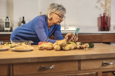 Olgun bir kadın sağlıklı bir yemek hazırlarken akıllı telefona odaklanır. Mutfakta, kırsal bir ahşap masada taze sebzeler ve malzemelerle çevrili.
