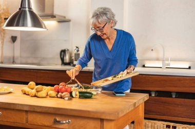 Mutlu, yaşlı bir kadın taze organik sebzelerle besleyici bir akşam yemeği pişiriyor. Sıcaklık ve neşe dolu, modern bir mutfakta emekliliğinin tadını çıkarıyor.