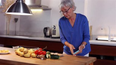 Üst düzey kadın sağlıklı yemek hazırlarken güvenli bir şekilde akıllı telefon kullanıyor. Dijital tarif rehberliğiyle modern mutfakta sebze doğruyor.