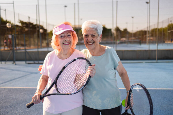 Two happy senior women are embracing and holding tennis rackets on a blue paddle tennis court, enjoying their active retirement and the benefits of physical activity
