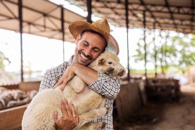 Neşeli çiftçi ahırın içinde bir kuzuyu nazikçe kucaklıyor, sürdürülebilir çiftlik hayvancılığında insanlar ve hayvanlar arasındaki iç açıcı bağı gösteriyor.