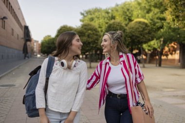 İki genç kadın, muhtemelen üniversite ya da lise öğrencisi, ağaçların oluşturduğu kampüs yolunda birlikte yürüyor, canlı sohbetler yapıyor ve birbirlerinin arkadaşlığından keyif alıyorlar.