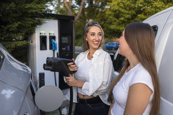 Two women charging their electric car at a station, smiling and engaging in conversation while holding the charging cable, embodying a modern and eco friendly lifestyle