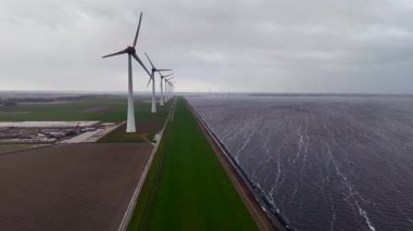 Kıyıdaki bir hendekte dönen uzun rüzgar türbinlerinin etkileyici hava perspektifi, bir tarafında deniz, diğer tarafında tarımsal alanlar olan sürdürülebilir elektrik üretiyor.