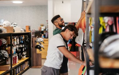 Dost canlısı bisikletçi çalışanı, genç bir erkek müşteriye raftan yeni bisiklet kıyafetleri seçerek yardımcı oluyor, perakende satış hizmeti sunuyor ve modern mağaza ortamında yararlı tavsiyelerde bulunuyor.