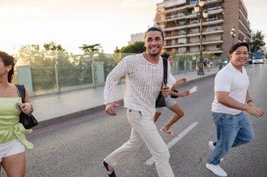 Bir grup mutlu çok ırklı arkadaş, şehir gecesinin tadını çıkarıyorlar, birlikte gülüyorlar ve arka planda binalar ve parlak bir günbatımı gökyüzü ile birlikte şehir caddesinde koşuyorlar.
