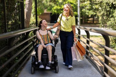 Anne ve yetişkin kız birlikte yürürken gülümsüyor ve gülüyorlar. Annesi alışveriş torbaları taşıyor ve elini kızının omzuna koyuyor.