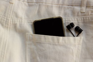 Siyah ve boş ekranlı cep telefonu ve arka cebinde beyaz kot pantolon olan kulaklıklar.