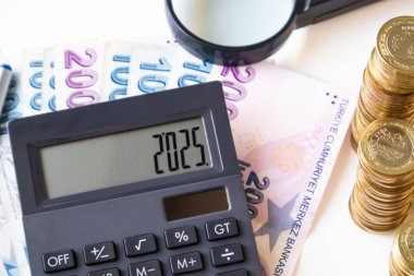 Türk Para Banknotları beyaz yüzey üzerinde madeni paralar ve üzerinde 2025 rakam bulunan siyah hesap makinesi
