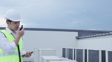 Asyalı bakım mühendisi, fabrikanın çatısındaki işi açıklamak için telsiz kullanıyor. Müteahhit kompresör sistemini denetleyecek ve modern binaların inşaat sahasına hava durumu sistemleri kurmayı planlıyor.