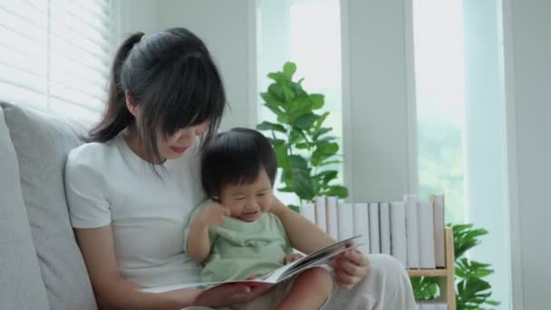 幸せなアジアの母は家で赤ん坊の時間と一緒に本をリラックスし 親は娘とソファに座って物語を読む ストーリーテリング 実践を学び — ストック動画