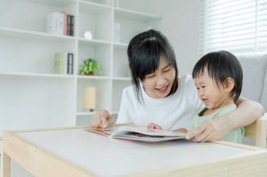 Mutlu Asyalı anne rahatla ve evde bebek zamanlarıyla kitap oku. Ebeveyn kızıyla kanepede oturup masal okuyor. öğrenme, çocuk bakımı, gülme, eğitim, hikaye anlatma, uygulama.