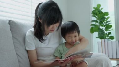 Mutlu Asyalı anne rahatla ve evde bebek zamanlarıyla kitap oku. Ebeveyn kızıyla kanepede oturup masal okuyor. öğrenme, çocuk bakımı, gülme, eğitim, hikaye anlatma, uygulama.