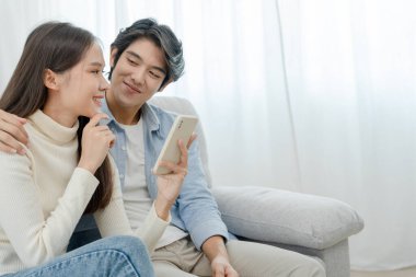 Gülümseyen Asyalı çift internet alışverişinde akıllı telefon kullanıyor. Koca, güzel bir eş için doğum günü hediyesi ödemeye hazır. Balayı için internetten konaklama, uygulama, uçak biletleri..