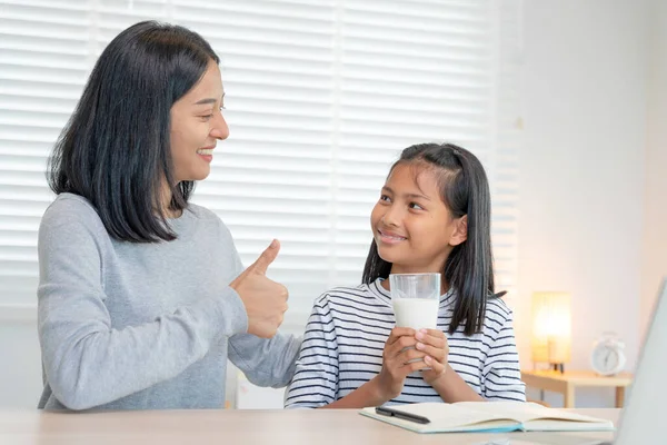 Asian young little girl learn at home. Do homework with kind mother help, encourage for exam. Mom pass on a glass of milk to daughter. Girl happy Homeschool. Mom teach and advice education together.