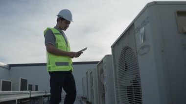 Asian maintenance engineer works on the roof of factory. contractor inspect compressor system and plans installation of air condition systems in construction. technology, online checking, mobile application.