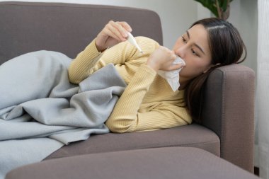 Yüksek ateşi olan genç bir Asyalı kadın vücut ısısını kontrol ederken, kadın hapşırması ve mevsimsel gribi olan akışkan burun, alerji, dijital termometre, virüs, koronavirüs, hastalık, solunum