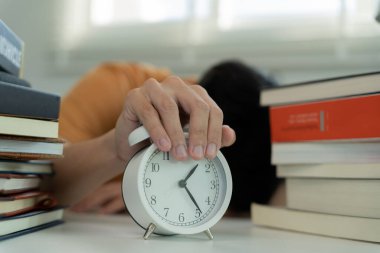Asian student man turns off the alarm clock because he is tired from reading, male prepare for test and learning lessons in the library. stress, despair, haste, misunderstanding reading, discouraged.