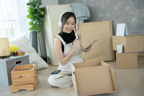 Move house, relocation. Woman using household checklist for new apartment Inside the room are cardboard boxes containing personal belongings and furniture. Move into a house or condominium
