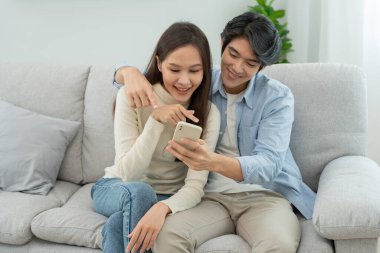 Gülümseyen Asyalı çift internet alışverişinde akıllı telefon kullanıyor. Koca, güzel bir eş için doğum günü hediyesi ödemeye hazır. Balayı için internetten konaklama, uygulama, uçak biletleri..