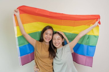 LGBT grubu. Güzel görünümlü lezbiyen çiftler gökkuşağı bayraklarını parlak bir şekilde örterler. Asyalı genç çift birbirlerine mutlu bir şekilde sarılıyorlar, aşık olan sevgili, biseksüeller, homoseksüellik, özgürlük, ifade, mutlu hayat.