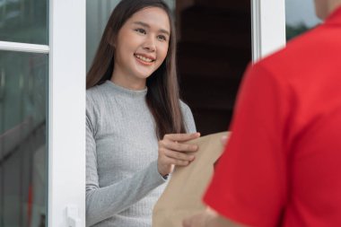Mutlu gülümseyen Asyalı kadın, kurye evinden bir paket yiyecek alıyor. Teslimatçı ekspres teslimat gönderdi. Çevrimiçi alışveriş, kağıt konteynır, paket servisi, postacı, dağıtım servisi, paketler