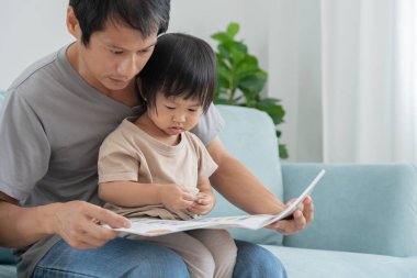 Mutlu Asyalı baba rahatla ve evde bebek zamanlarıyla kitap oku. Ebeveyn kızıyla kanepede oturup masal okuyor. öğrenme, çocuk bakımı, gülme, eğitim, hikaye anlatma, uygulama.