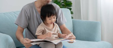 Asyalı baba evde bebek zamanı ile kitap okuyup rahatlıyor. Ebeveyn kızıyla kanepede oturup masal okuyor. öğrenme, çocuk bakımı, gülme, eğitim, hikaye anlatma, uygulama.