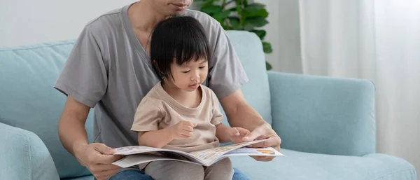 Asian father relax and read book with baby time together at home. parent sit on sofa with daughter and reading a story. learn development, childcare, laughing, education, storytelling, practice.