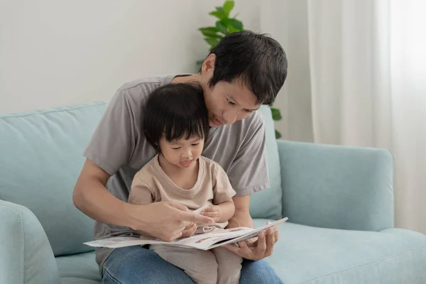 Happy Asian father relax and read book with baby time together at home. parent sit on sofa with daughter and reading a story. learn development, childcare, laughing, education, storytelling, practice.