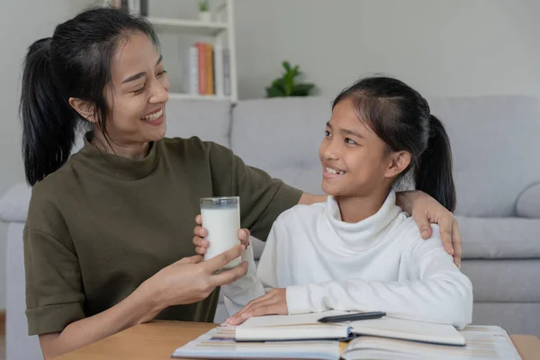 Asian young little girl learn at home. Do homework with kind mother help, encourage for exam. Mom pass on a glass of milk to daughter. Girl happy Homeschool. Mom teach and advice education together