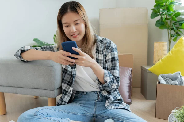 Moving house, relocation. asian woman feel good and recreation and use phone on new house, inside the room was a cardboard box containing personal belongings and furniture. move in condominium