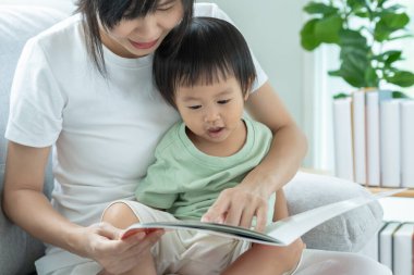 Mutlu Asyalı anne rahatla ve evde bebek zamanlarıyla kitap oku. Ebeveyn kızıyla kanepede oturup masal okuyor. öğrenme, çocuk bakımı, gülme, eğitim, hikaye anlatma, uygulama.