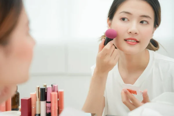 Beautiful Asian woman sit in front of a mirror and smile on makeup. face of a healthy woman applying makeup. Advertisement, lifestyle , cosmetics, makeup accessories, beauty activity, beautician