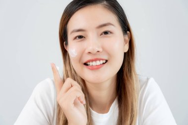 Güzel Asyalı kadın gülümsüyor, cilt kremi kullanıyor. Sağlıklı bir kadın yüzüne krem sürün ve makyaj yapın. Cilt kremi reklamı, kırışıklık önleyici, bebek yüzü, beyazlatma, nemlendirici, gözenekleri sıkılaştırma serumu.