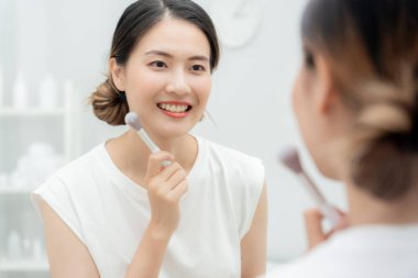 Güzel bir Asyalı kadın aynanın karşısına oturur ve makyaja gülümser. Makyaj yapan sağlıklı bir kadının yüzü. Reklam, yaşam tarzı, kozmetik, makyaj malzemeleri, güzellik aktiviteleri, güzellik