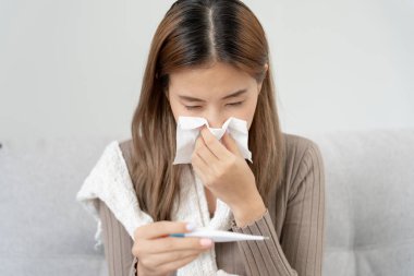 Young Asian woman having high fever while checking body temperature, female sneezing and runny nose with seasonal influenza, allergic, digital thermometer, virus, coronavirus, illness, respiratory