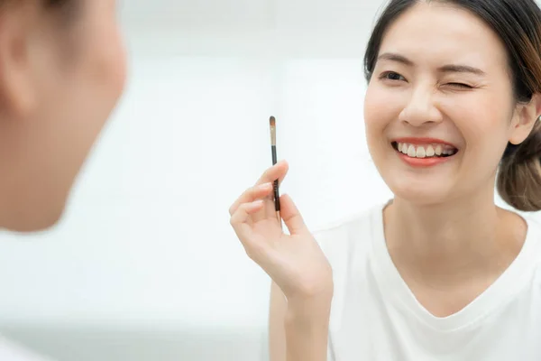 Beautiful Asian woman sit in front of a mirror and smile on makeup. face of a healthy woman applying makeup. Advertisement, lifestyle , cosmetics, makeup accessories, beauty activity, beautician