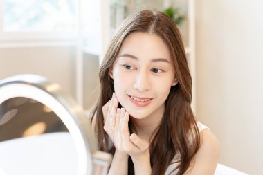 Güzel bir Asyalı kadın aynanın önünde oturur. Krem sürerek makyaj yapan sağlıklı bir kadın yüzü. Cilt kremi reklamı, kırışıklık önleyici, bebek yüzü, beyazlatma, nemlendirici, gözenekleri sıkılaştırma serumu.