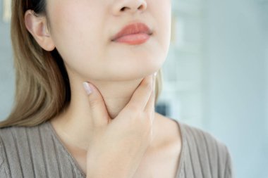 Asyalı kadın boğazı ağrıdığı için elini boynuna koydu. Dişi bademcik iltihabı iltihaba neden olur. Su alarak, antibiyotik alarak, doktora, virüse, korona virüsüne, gribe, gribe danışarak tedavi olunur.