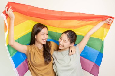 LGBT grubu. Güzel görünümlü lezbiyen çiftler gökkuşağı bayraklarını parlak bir şekilde örterler. Asyalı genç çift birbirlerine mutlu bir şekilde sarılıyorlar, aşık olan sevgili, biseksüeller, homoseksüellik, özgürlük, ifade, mutlu hayat.