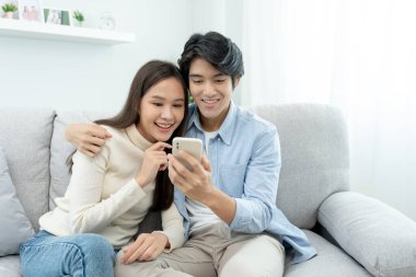 Gülümseyen Asyalı çift internet alışverişinde akıllı telefon kullanıyor. Koca, güzel bir eş için doğum günü hediyesi ödemeye hazır. Balayı için internetten konaklama, uygulama, uçak biletleri..