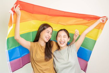 LGBT grubu. Güzel görünümlü lezbiyen çiftler gökkuşağı bayraklarını parlak bir şekilde örterler. Asyalı genç çift birbirlerine mutlu bir şekilde sarılıyorlar, aşık olan sevgili, biseksüeller, homoseksüellik, özgürlük, ifade, mutlu hayat.