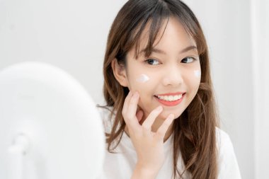 Güzel bir Asyalı kadın aynanın önünde oturur. Krem sürerek makyaj yapan sağlıklı bir kadın yüzü. Cilt kremi reklamı, kırışıklık önleyici, bebek yüzü, beyazlatma, nemlendirici, gözenekleri sıkma seru