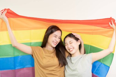 LGBT grubu. Güzel görünümlü lezbiyen çiftler gökkuşağı bayraklarını parlak bir şekilde örterler. Asyalı genç çift birbirlerine mutlu bir şekilde sarılıyorlar, aşık olan sevgili, biseksüeller, homoseksüellik, özgürlük, ifade, mutlu hayat.