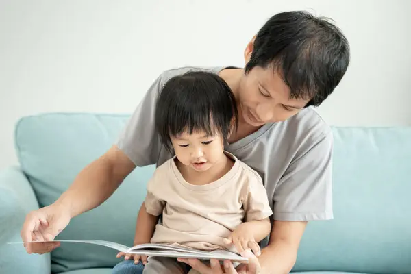 Happy Asian father relax and read book with baby time together at home. parent sit on sofa with daughter and reading a story. learn development, childcare, laughing, education, storytelling, practice