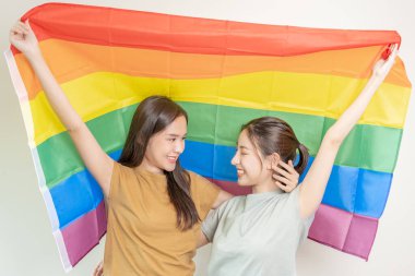 LGBT grubu. Güzel görünümlü lezbiyen çiftler gökkuşağı bayraklarını parlak bir şekilde örterler. Asyalı genç çift birbirlerine mutlu bir şekilde sarılıyorlar, aşık, biseksüellik, homoseksüellik, özgürlük, ifade, mutlu bir hayat.
