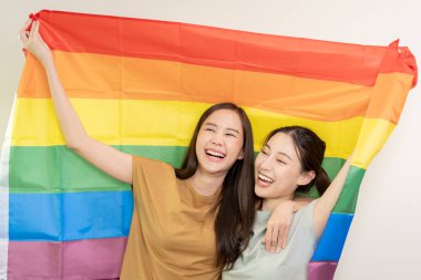 LGBT grubu. Güzel görünümlü lezbiyen çiftler gökkuşağı bayraklarını parlak bir şekilde örterler. Asyalı genç çift birbirlerine mutlu bir şekilde sarılıyorlar, aşık olan sevgili, biseksüeller, homoseksüellik, özgürlük, ifade, mutlu hayat.